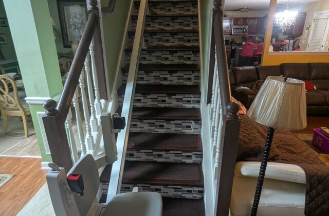 Bruno Elan stairlift at the foot of a carpeted staircase beside a living room lamp