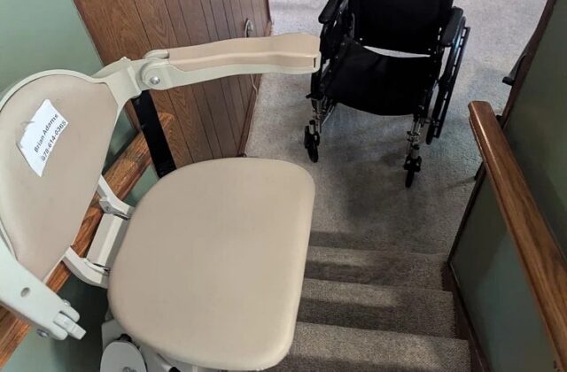 Bruno stairlift at the top of carpeted stairs, a wheelchair parked in the room beyond