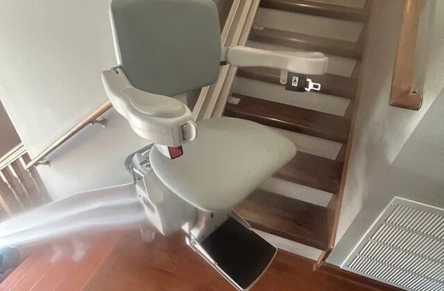 Bruno stairlift with a grey seat at the foot of carpeted stairs beside a floor vent