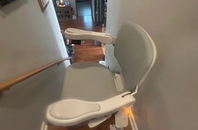 Bruno stairlift parked at the top of stairs in a hallway, seat folded and arms raised