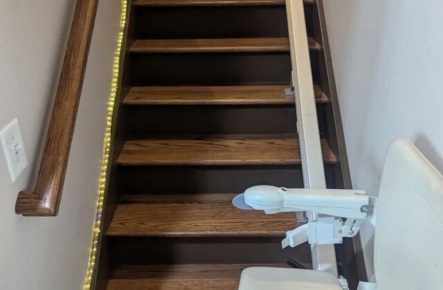 Handicare stairlift parked at the top of a narrow wooden staircase beside a handrail