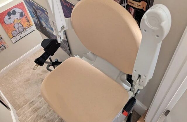 Handicare stairlift with a beige seat at the top of carpeted stairs, framed art on the wall