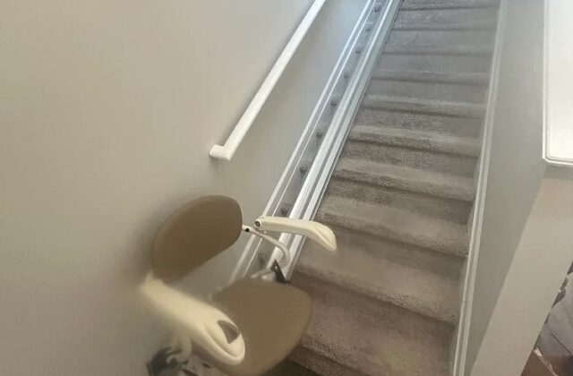 Harmar stairlift at the foot of a long carpeted staircase with its rail rising to the top