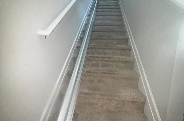 Harmar stairlift rail running up a long carpeted staircase beside a white handrail