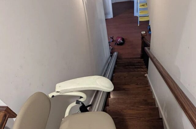 Harmar stairlift with a beige seat and seat belt at the top of narrow wooden stairs