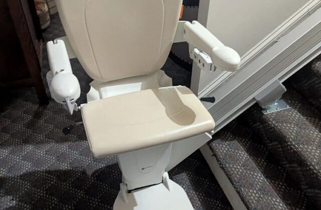 Stairlift with a cream seat at the foot of carpeted stairs in a living room