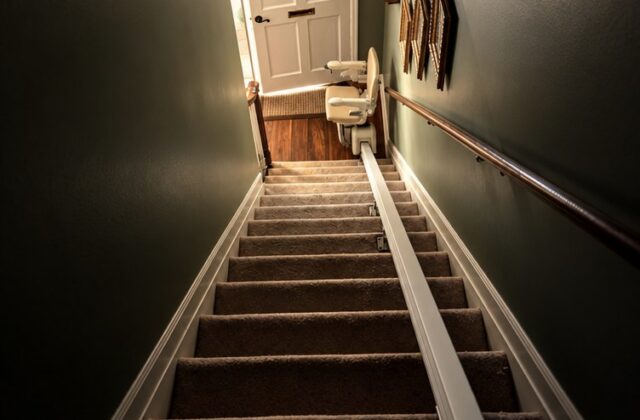 Stairlift rail running down carpeted stairs in a dark-walled stairwell toward a front door