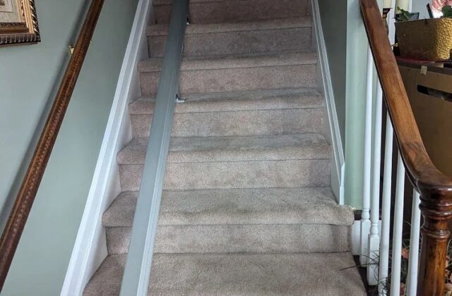 Stairlift rail along carpeted stairs next to a wooden banister, chair folded at the bottom