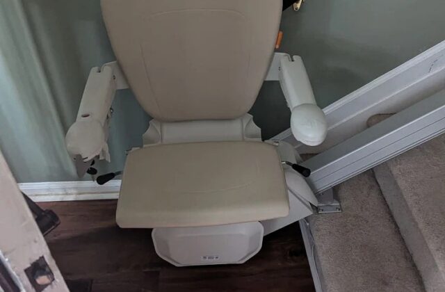 Stairlift with a beige seat parked at the top of stairs beside a wooden handrail