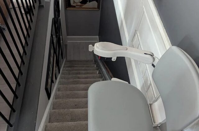 Bruno stairlift with a grey seat at the top of a carpeted staircase beside a black iron railing, on a service visit